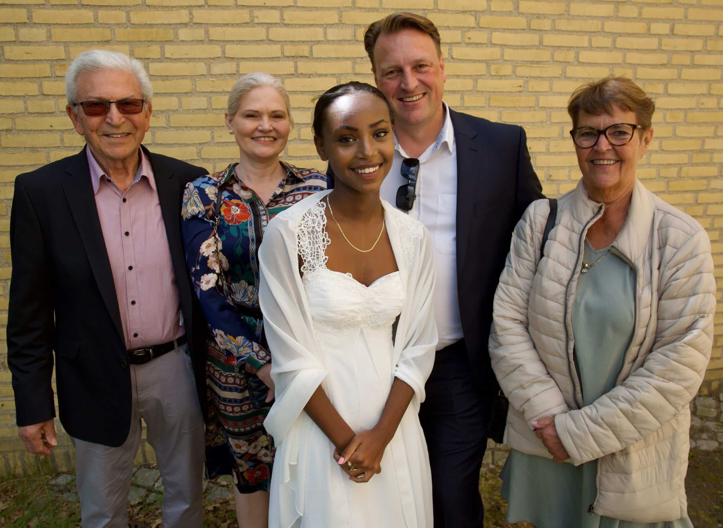 Konfirmationsfotografernne har her fotograferet en familie foran en gul murstenskirke. Billedet viser den kvindelige konfirmand med hendes forældre og bedsteforældre