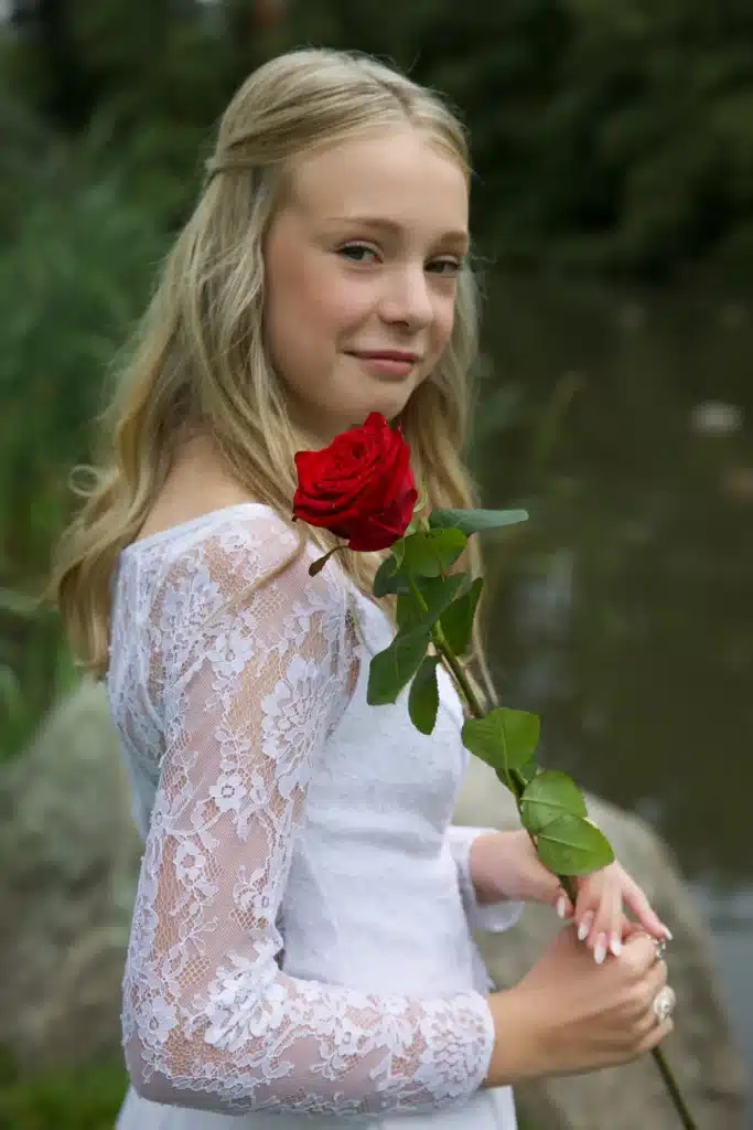 Kvindelig konfirmand i blondekjole står med langstilket rød rose i hånden