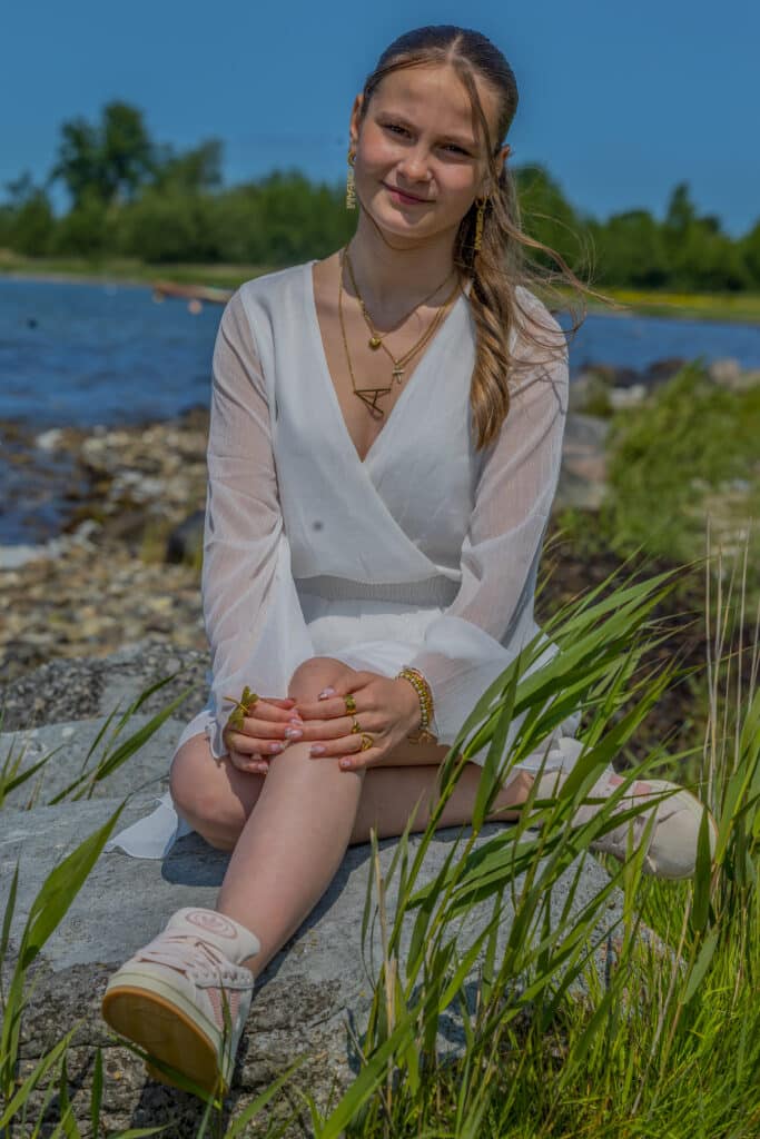 Kvindelig konfirmand sidder på en sten og poserer med vand i baggrunden