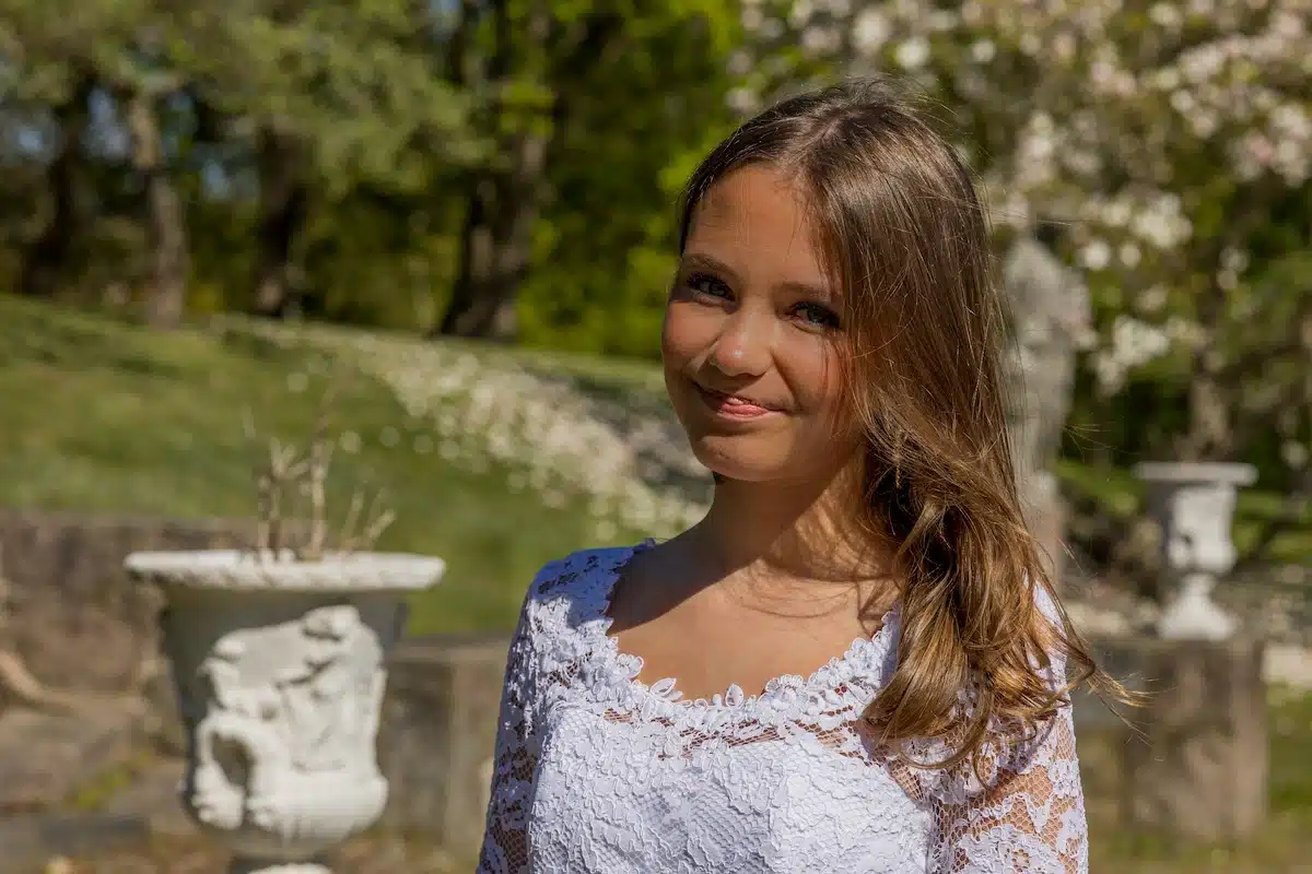 Festfotograferne har her været ude til en konfirmation af tvillingepiger. På billedet ses den ene tvilling i sin hvis blondekjole. I baggrunden ses en have med hvide marmorskulptur samt blomstertræer med hvide blomster
