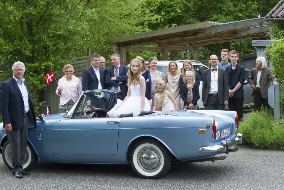 Festfotograferne har her fotograferet en kvindelig konfirmand, der sidder i en lyseblå sportsvogn og med familien der står i baggrunden