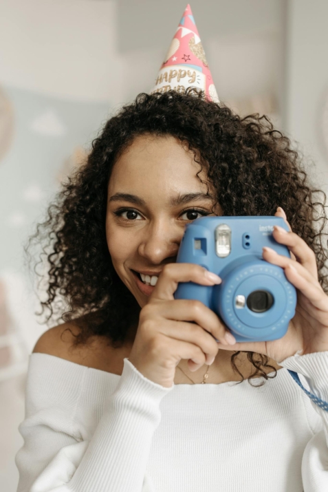 A cheerful woman with curly hair and a party hat holding a blue polaroid camera indoors.