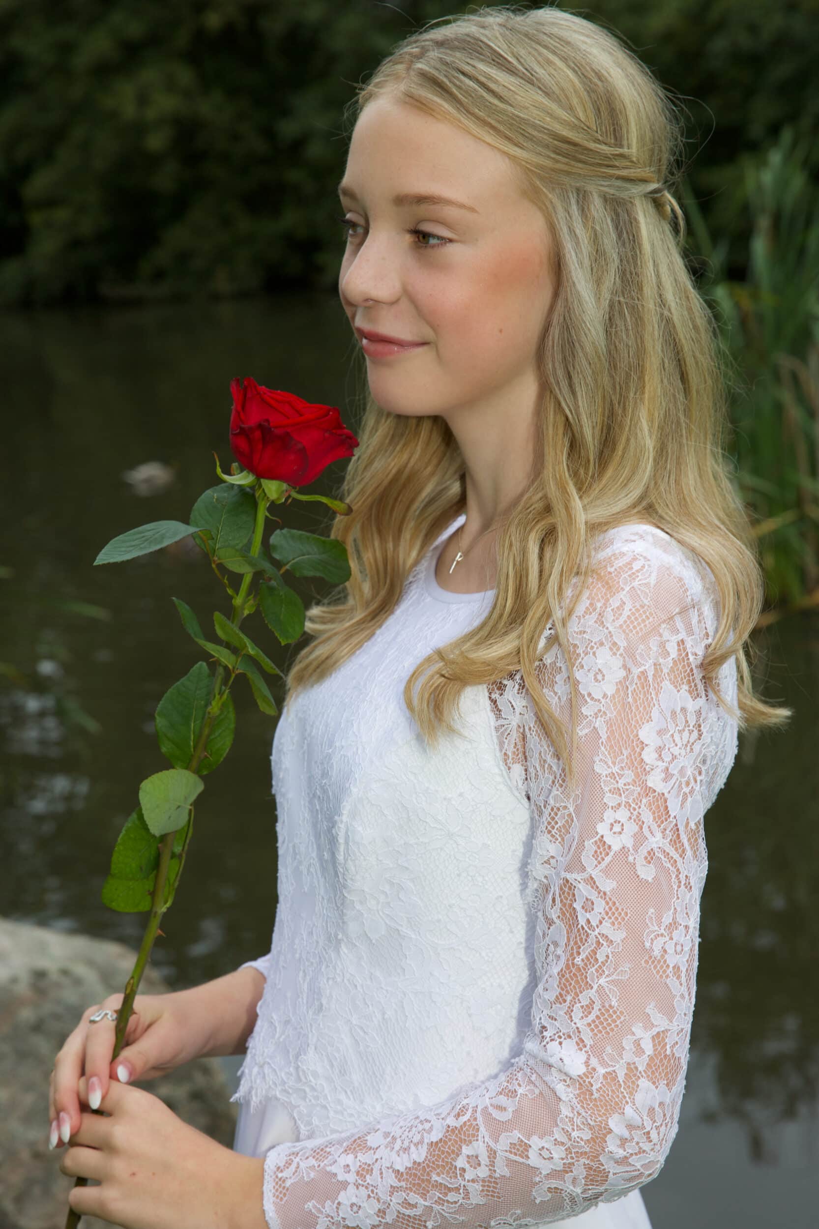 Festfotografen har her fotograferet en kvindelig konfirmand i blondekjole. Konfirmanden ses fra siden og holder en rød langstilket rose i hånden